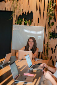 Johanna Voss, fractional COO and talent management agency consultant, smiling during a working session at a co-working space desk with her laptop open, surrounded by colleagues